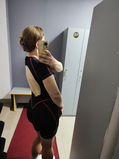 A woman is taking a selfie in a locker room. She is wearing a black swimsuit with pink trim and has her hair pulled back in a bun with a large clip. She is holding a gold smartphone up to take the photo. In the background, there are gray lockers, a bench with a yellow stripe, and a red carpet runner. The floor is tiled. The scene appears to be in a public facility, possibly a gym or sports club, in Korosten, Ukraine.