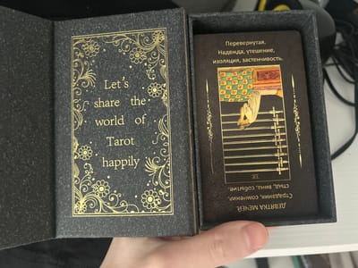 An open, dark gray, fabric-covered box containing two sides. The left side of the box opens like a book and displays a textured, dark gray surface with a decorative gold border. Inside this border, text in gold lettering reads "Let's share the world of Tarot happily." The right side of the box holds a deck of tarot cards. The top card is visible, showing an illustration with a green and red checkered pattern at the top, a person in a white garment, and multiple swords arranged horizontally below. The text on this card is in Russian and appears to be upside down. The card's title is "ДЕВЯТКА МЕЧЕЙ", which translates to "Nine of Swords". The text above the illustration reads "Перевернутая. Надежда, утешение, изоляция, застенчивость." (Reversed. Hope, consolation, isolation, shyness.). The background of the card is dark brown with gold decorative elements. The overall scene is indoors with a white surface visible in the foreground where a hand is holding the box open. The image's geolocation is specified as Bugry, Russia.