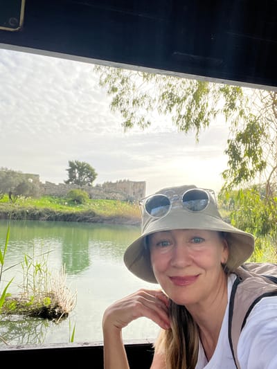 The image depicts an outdoor scene in Rosh Haayin, Israel, featuring a woman in the foreground with the Yarkon River and Antipatris Fortress in the background.

A middle-aged woman, with light skin and light brown hair, occupies the lower right portion of the frame. She is smiling faintly and looking directly at the camera. She wears a light khaki-colored bucket hat with clear-framed sunglasses perched on top. Her left hand is resting on a dark horizontal surface, possibly a railing. A white garment is visible under a light-colored backpack strap on her right shoulder.

Behind the woman, the Yarkon River flows horizontally across the middle ground. Its water is a greenish-blue, reflecting light. Reeds and other vegetation line the riverbanks and form small clusters within the water. On the far bank, partially obscured by greenery, are the stone ruins of a large structure, identified as the Antipatris Fortress. The fortress walls are weathered and appear to be constructed from light-colored stone, with some openings visible. Trees and green grass surround the ruins.

The sky above is partially cloudy with scattered white clouds against a lighter blue, suggesting an overcast or partly cloudy day. A bright spot, indicative of the sun, is visible in the upper right quadrant. A tree with light green foliage hangs into the frame from the top right. A dark, solid horizontal beam or structure runs across the top of the image, and a similar dark horizontal surface is present at the bottom, suggesting the vantage point is from within a covered viewing area or structure.