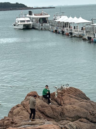 The image displays a coastal scene within Shinan District, China, under an overcast sky. In the foreground, a large, textured reddish-brown rock formation is present. Two individuals are on these rocks: a female in a green long-sleeved top and dark trousers is seated higher on the right, facing left with a slight smile; a male in a light brown jacket and dark trousers stands lower on the left, facing partially away from the viewer with his head angled downwards. A thin rope or cable extends horizontally across the midground, between the rocks and the water. In the background, calm grey-blue water extends to the horizon. Several boats are moored to a floating dock structure: a white catamaran-style yacht is partially visible on the left, and a series of at least four flat-decked recreational boats, each topped with white tent-like awnings, are arranged in a line to the right. Distant landmasses and faint vessel outlines are visible on the horizon.