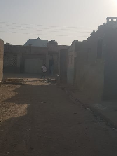 The image depicts a street scene in Dutsinma B, Nigeria. The dirt street runs along a row of buildings of varying heights and designs, constructed of aged materials. Two people are visible on the street. One person, likely a male, is wearing a light-colored shirt and dark pants, and appears to be standing next to a bicycle. The second person is standing close to the first person. Overhead power lines are visible in the sky. The image is taken in daylight, with a hazy or smoky atmosphere.