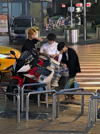 The image depicts a street scene at night in the city of Longta, China, possibly near a crosswalk or public area with bike racks. Three young individuals are the primary focus, gathered around a parked motorcycle and a scooter.
On the left, a person with blonde hair and a black shirt is leaning forward, with their right arm bent and hand near their chest, seemingly engaged with their phone or a similar device. Next to them, in the center, a person wearing a white jacket is looking down at a mobile phone. To the right, a person in a black textured jacket and plaid pants is seated on the scooter, also looking downwards, with their hands resting on their lap or the vehicle.
The motorcycle, prominently featured in the foreground, is a red and white sportbike with the branding "SPORTS VR46" and "560" visible. A black helmet rests on the motorcycle's seat. The scooter is light blue and partially obscured.
The environment suggests a wet or recently rained-on street, with reflections visible on the pavement. The background shows a dark building with illuminated signage and some streetlights, contributing to the nighttime atmosphere. Other elements in the scene include a yellow vehicle to the left, a silver van further back, and metal bike racks in the foreground and middle ground. Construction cones are visible in the distance, suggesting ongoing work or temporary traffic management. The overall impression is a casual gathering of young people on a street in an urban setting.