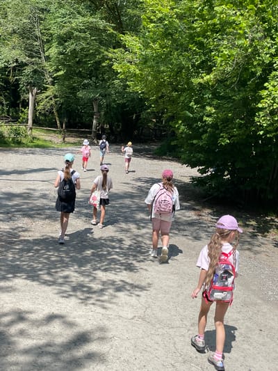 The image shows a group of children walking along a gravel path in a wooded area. The sun is shining, casting shadows of the trees onto the path. The children are wearing casual clothing and backpacks, suggesting they are on an outing or field trip. The scene is set in Bolshoy Sochi, Russia, a region known for its natural beauty and outdoor recreational opportunities. The overall mood appears to be lighthearted and adventurous, with the children seemingly enjoying their walk. No visible text is present in the image.