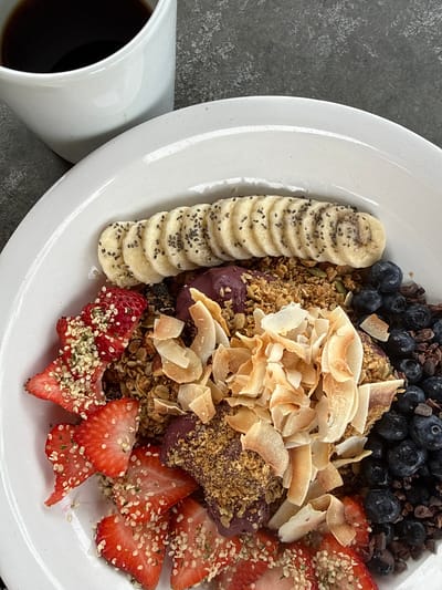 The image displays a healthy breakfast or brunch meal featuring an acai bowl and a cup of coffee.
**Subjects:** The main subject is a vibrant acai bowl filled with various colorful toppings, presented on a round, white ceramic plate. Alongside the plate, a simple white ceramic mug holds a dark liquid, which appears to be coffee.
**Activity or event taking place:** The image captures a moment of a prepared meal, ready to be consumed, suggesting a leisurely breakfast or brunch.
**Notable details:**
* **Acai Bowl Contents:** The acai base is a deep purple color, mostly covered by an abundant arrangement of toppings.
* Along the left edge, a neat line of sliced bananas is visible, each piece sprinkled with small black chia seeds.
* The upper right section features a generous portion of fresh blueberries, intermingled with dark, irregularly shaped pieces that resemble cacao nibs or dark chocolate.
* A significant amount of golden-brown granola, containing visible pumpkin seeds, forms a base for other toppings.
* Piled prominently in the center are light brown, flaky toasted coconut chips, adding texture and warmth.
* The lower right portion of the bowl is adorned with bright red, sliced strawberries, heavily sprinkled with light green or beige hemp seeds.
* **Coffee:** The white mug contains a dark, presumably black, coffee.
* **Setting:** The plate and mug rest on a dark gray, subtly textured surface, likely a stone countertop or table, which provides a neutral and modern backdrop.
* **Presentation:** The food is artfully arranged, emphasizing its freshness and visual appeal. The meal appears wholesome and nutritious.
* **Time of Day:** The nature of the meal suggests it is consumed in the morning or early afternoon.
**Visible text:** There is no visible text in the image.