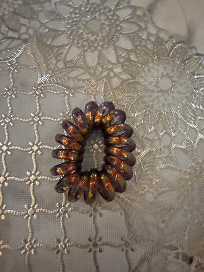 A coiled hair tie, iridescent purple and orange, rests on a white textured surface, possibly lace or a doily. The surface has a floral pattern with intricate stitching, and it appears to be part of a tablecloth or decorative placemat. The hair tie is the central object of focus, its spiral form catching the light. The background is softly blurred, emphasizing the texture and detail of the doily. The lighting is natural, casting subtle shadows and highlighting the reflective qualities of the hair tie. There are no people or animals present in the image. The scene appears to be indoors, likely on a table. The location within the city of City of Pematang Siantar, Indonesia is not determinable from the visual information provided.