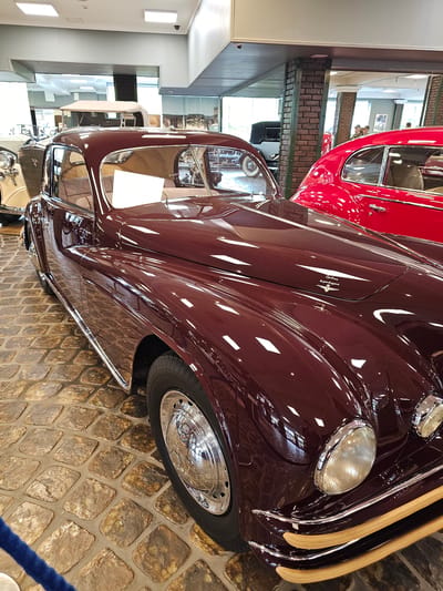 The image shows a close-up of two vintage cars in a museum setting. The primary car, in the foreground, is a maroon color with a highly polished finish, reflecting light and highlights. Its headlights are prominent, and a strip of wooden trim is visible along the front. The wheel features a chrome hubcap. The second car, in the background, is red. Both cars are parked on a cobblestone-like surface. A glimpse of the museum's interior is visible, with windows and architectural details suggesting the location might be in Архангельское, Russia, as museums are common in such historical places. The image is taken at a slight angle, giving a dynamic perspective of the cars.