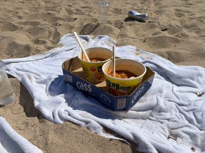 The image shows two containers of Ottogi Chamgae Ramen in a blue cardboard Cass beer box, placed on a white towel on a sandy beach. The containers have chopsticks in them, and the ramen appears to be prepared with broth and noodles visible. In the background, there is a discarded plastic cup and a crushed aluminum can on the sand. To the left, a water bottle is partially visible. The sand texture suggests a natural beach environment. Based on the Ottogi and Cass brands, along with the ramen packaging, this is likely in South Korea, potentially near the city of Toseong-myeon.