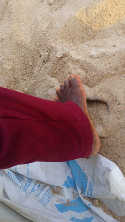 A person's bare foot and lower leg, clad in maroon pants, rest on a white and blue patterned sack partially filled with sand. The scene is set on a sandy surface, presumably a beach or similar environment. The location is implied to be near Eziukwu, Aba, Nigeria, based on text visible on the sack.