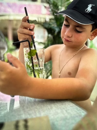 The image shows a young boy wearing a black baseball cap with a white horse logo. He is shirtless and has a gold chain around his neck. He is holding a glass with a drink that appears to be a mojito, complete with ice, mint leaves, and lime slices. Two black straws are in the glass. The boy is looking down at the drink with a relaxed expression. The background is slightly blurred, showing greenery and what appears to be outdoor seating with a light-colored surface. The overall lighting suggests it is daytime, possibly outdoors in a casual setting like a cafe or patio. The image was taken in Artik, Armenia.