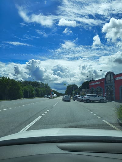 The image shows a view from a car on a road in Yessentuki, Russia, with a large red building on the right side of the road. The building has arched windows and signage that reads "ГОРЯЧИЕ ПИРОЖКИ". Several cars are parked in front of the building, and a few people are visible near the entrance. The road has multiple lanes and white dashed lines marking the lanes. On the left side of the road, there are trees and more cars driving. The sky is blue with large, fluffy white clouds, indicating a sunny day. The image appears to be taken during daylight hours.