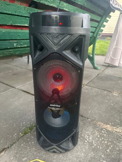 This media file features a black portable speaker resting on a gray concrete patio. The speaker has a mesh grill through which two illuminated drivers are visible; one emits a red light, and the other displays a blue and yellow pattern. The "smartbuy" logo is visible on the mesh grill. To the left of the speaker, a portion of a green wooden bench with peeling paint is visible, and beyond that, a white tarp is partially visible, along with some greenery and a building. The scene appears to be outdoors, possibly in a yard or garden. The lighting suggests it is daytime, and the concrete surface shows some signs of wear and tear, with a small amount of dried grass and dirt. No people are present, and the overall atmosphere is neutral.
