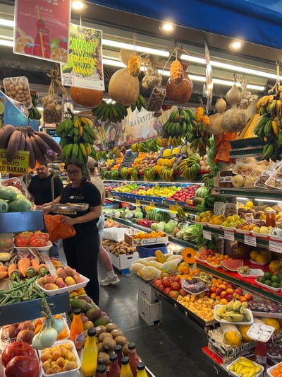 Here is a description of the image:
The image shows a vibrant, bustling fruit and vegetable market in Las Palmas, Spain. The scene is filled with a colorful array of fruits and vegetables, meticulously arranged in stalls. Bananas, melons, tomatoes, peaches, and other produce are abundantly displayed.
People are present, including a woman in a black shirt wearing glasses, who appears to be weighing produce. Another individual, also wearing a black shirt, is nearby. There are other people in the background, including one person wearing light colored shorts and white sneakers.
Signs with descriptions and prices are visible throughout the market, including "CANARY ECOLOGICAL BANANA" and "ROJO RED BANANA." The image has a slight tilt, giving the impression of looking upward or downward along the length of the market stall. Artificial lights are visible on the ceiling of the marketplace.