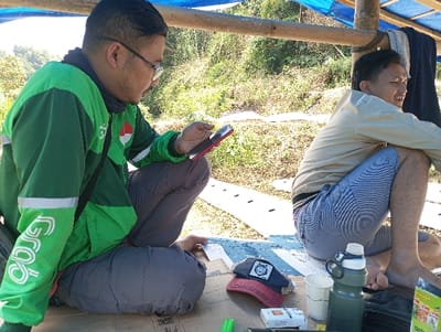 Two males are present under a makeshift shelter in an outdoor, vegetated environment within Cibeureum, Indonesia. The shelter consists of bamboo poles supporting a blue tarp. Both individuals are seated on a flat, light-colored surface, possibly cardboard or a mat.
The male to the left, wearing a dark green Grab-branded jacket with white accents, grey trousers, and eyeglasses, holds a red-cased smartphone in his right hand, looking down at the device.
The male to the right is barefoot, wearing a light-colored jacket and blue-and-white striped shorts. He is in a deep squatting posture, facing to his right, with his attention directed off-camera.
On the surface between them are a dark blue baseball cap with a white emblem, a red baseball cap, a white Marlboro cigarette pack, a dark green water bottle with a white cap, and two white disposable cups. The background reveals a sun-drenched hillside covered with sparse green and brown vegetation, featuring visible linear ground markings in the mid-distance.