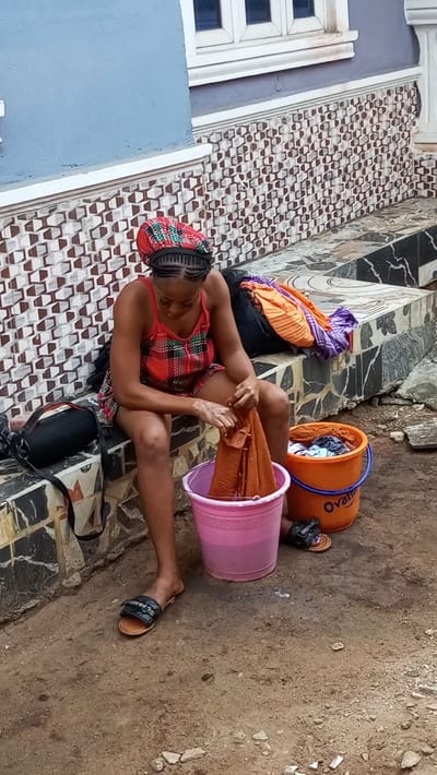 In Agburu Uke, Nigeria, a young woman is depicted hand-washing clothes outdoors during the daytime. She is seated on a low, tiled ledge, with her head bent in concentration over a task. She wears a vibrant, patterned headwrap over her braided hair, a red and green plaid camisole top, and red shorts. Black slides, one with "Supreme" visible, are on her feet.
In front of her is a pink bucket filled with water, where she is actively wringing or scrubbing a piece of orange/brown fabric. To her right, an orange bucket contains more clothes, with the brand name "Ovaltine" clearly visible on its side. Another pile of folded clothes, including orange and purple garments, rests on the tiled ledge behind the orange bucket. A black bag with a strap is placed on the ledge to her left.
The setting is against a building featuring a blue-painted upper wall and a lower section adorned with intricate, geometric brown and white tiles. A white window frame is visible on the blue wall. The ground beneath her is bare earth, suggesting a common outdoor area. The lighting indicates a clear day. The woman appears focused and diligent in her activity.