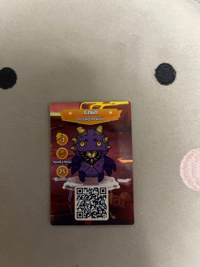 The image shows a close-up of a collectible card featuring a cartoonish, purple monster with yellow eyes and horns. The card has text in Cyrillic that translates to "Alp, Legendary" at the top. Below the monster, there are icons and text indicating a power level of "3" and a number "25" labeled "number." A QR code is also visible on the card. The card is placed on a soft, textured surface, likely a blanket or pillow, which appears to be light gray with two dark circular spots. There are no visible elements in the image that could be used to determine its location in Berezhany, Ukraine.