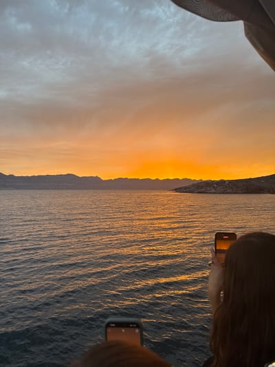 A horizontal image captures a scene during sunset with a body of water, possibly an ocean or a large lake, occupying the right half. The water reflects the warm hues of the setting sun, creating a shimmering effect. The left half of the image features the sky transitioning from soft, warm colors near the horizon to cooler tones higher up. A silhouette of a landmass, potentially a mountain range or coastline, separates the sky from the water.
Two people are visible, each holding a smartphone. The first person on the upper right side is actively filming or taking a photo, as indicated by the recording icon on their phone screen. The second person is located on the lower right, also holding a smartphone.
A sheer curtain hangs in the upper-left corner of the frame, suggesting the photograph was taken from inside a building or structure.
The location is Unknown, Mexico.