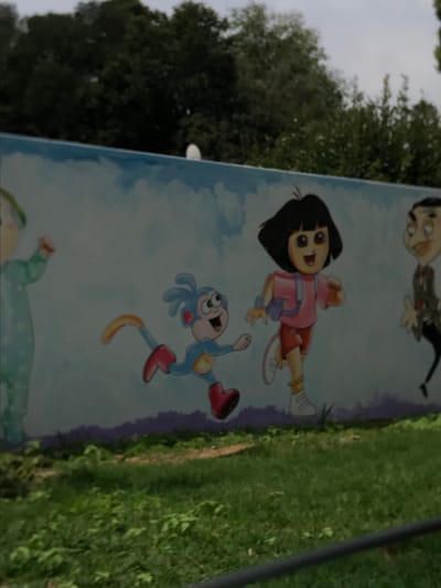 A painted mural is depicted on a light-colored wall in Germiston, South Africa. The mural features multiple cartoon characters against a sky-blue background with white clouds and a purple ground line. From left to right, a character in light blue pajamas with a green cap is partially visible, presenting a raised right arm. Adjacent to this, Boots the monkey, rendered in blue fur with red boots and a yellow tail, is illustrated in a running pose. To the right, Dora the Explorer, wearing a pink shirt, orange shorts, white sneakers, and a pink backpack, is also depicted in a running posture. A partial rendition of Mr. Bean is visible on the far right. Above the mural wall, dense green trees and foliage are observed beneath an overcast sky. The foreground consists of green grass and weeds, with a dark horizontal bar crossing the bottom of the frame.