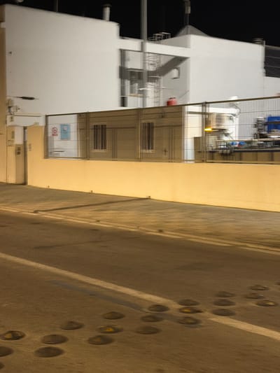 The image depicts an industrial or utility facility at night. In the background, a large, white, multi-story building dominates the scene, featuring various structural elements like staircases and pipes. A chain-link fence runs across the middle ground, separating the foreground from the building's immediate surroundings. Beyond the fence, glimpses of industrial equipment, including tanks and machinery, are visible.

The foreground consists of a paved area, likely a road or walkway, illuminated by artificial lights. This area features a pattern of raised circular markers embedded in the surface, possibly for traffic guidance or to delineate parking spaces. The overall lighting suggests it is nighttime, with artificial lights casting shadows. The environment appears to be an outdoor industrial zone.

Regarding the location in Ibiza, Spain, while the image itself does not contain explicit identifiers, the presence of industrial facilities at night, especially in a tourist destination like Ibiza, could be consistent with infrastructure such as a power plant, water treatment facility, or a port-related service area. Without further context, pinpointing the exact location within Ibiza is not possible based solely on the visual information.
