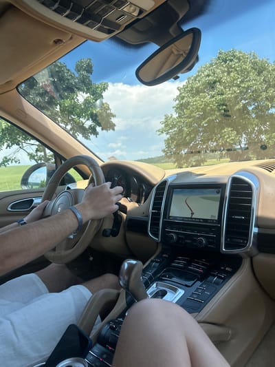 The image depicts the interior of a tan-colored Porsche Cayenne, observed from the front passenger seat, in the vicinity of Bliznatsi, Bulgaria. A driver, identified by forearms and hands, steers the vehicle. The driver wears white shorts, a blue wristband on the left wrist, and a silver wristband on the right wrist with partially visible text "NATIONAL". The driver's hands are positioned on the tan, leather-wrapped steering wheel, which displays the Porsche crest.
The center console features a prominent infotainment screen displaying a navigation map with a red route line, indicating a turn in 50 meters, and a time stamp of "13:17 TP". Below the map, "AUX BT - Suave (Kiss Me) (feat. Me..." indicates audio playback. The screen is flanked by two chrome-trimmed air vents. Below the screen, the climate control panel shows a temperature setting of "18.0" (likely Celsius) and an "AUTO" button engaged. A tan automatic gear shifter is visible on the center console.
In front of the passenger, a dark-colored smartphone with a blue case and a silver beverage can rest on the console. The passenger's knees/lower thighs, clad in light-colored attire, are visible in the foreground.
Through the windshield, a clear blue sky with scattered white clouds is visible. Lush green deciduous trees with dense foliage are prominent, indicating a wooded or rural environment. Rolling green fields are visible in the background, particularly on the left side. A faint, light-colored building with a dark roof is discernible in the distant landscape. The interior rearview mirror, also tan, is affixed to the windshield. The overall lighting suggests a bright, sunny day.