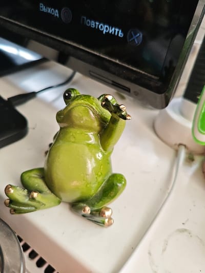 A detailed, close-up, slightly high-angle shot captures a glossy green frog figurine as its central subject. The figurine, appearing to be ceramic or resin, features varied shades of green, with darker mottling and yellowish highlights on its belly. Its prominent black eyes feature distinct white reflections. The frog's front right limb is raised, fingers splayed, and all visible digits on its limbs are tipped with a metallic gold finish.
The figurine rests on a flat, white, slightly dust-laden surface, which exhibits minor smudges and faint circular residue marks. In the upper background, a black electronic screen displays two illuminated Russian words: "Выход" (Exit) on the left, accompanied by a circular symbol, and "Повторить" (Repeat) on the right, next to an "X" symbol. The screen's bezel and the top of the device also show a thin layer of dust. Bright white reflections from an unseen light source are visible on the screen's surface and the frog's head. A black cable is partially visible to the left of the frog, while a thinner white cable connects to a white and green electronic device, partially obscured on the right side of the white surface. The bottom-left corner reveals a portion of a metallic edge and a black gridded pattern. There are no identifiable features within the image that indicate the location is Mława, Poland.