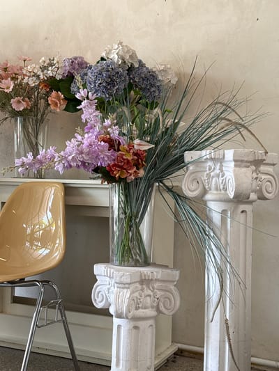 A still life composition features several arrangements of artificial flowers and decorative architectural elements against a textured, light-colored wall.
In the foreground to the left, a glossy, mustard-yellow chair with a chrome frame is partially visible. The chair reflects light, showing hints of an outdoor scene.
The main focus is on multiple vases of artificial flowers. One vase, placed on a white pedestal with Corinthian-style carvings, holds a large bouquet featuring prominent blue and purple hydrangeas, pale pink and white flowers, and some orange-red blooms. Long, thin green grass-like stalks emerge from this arrangement and arch across the frame.
To the left of this main bouquet, another glass vase holds a different arrangement of lighter pink and white flowers, with some taller, more delicate blooms in a pale purple hue extending from it. Behind these, a third vase is visible, containing dried, grass-like stems.
To the right of the central floral arrangement, two white decorative columns with Corinthian-style capitals are positioned. The column on the right is taller and more fully visible than the one on the left, which is partially obscured by the flowers and the other column.
The background is a plain, textured wall with subtle imperfections. The overall lighting is soft, suggesting an indoor setting.
This image does not provide any specific information to suggest its location in Tam Kỳ, Vietnam.