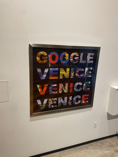 A framed artwork hangs on a white wall. The artwork features the word "GOOGLE" in the top row, followed by "VENICE" in the second row, "VENICE" in the third row, and "VENICE" in the fourth row. Each letter within these words is filled with a colorful, abstract image or pattern. The frame is metallic and appears to be silver or brushed nickel. To the left of the artwork, there is a white square panel on the wall, possibly an electrical panel or access point. To the right of the artwork, a portion of a white cabinet or fixture is visible. The wall meets the floor with a black baseboard. The floor is concrete with visible aggregate. An electrical outlet is visible on the wall below the right side of the artwork. The overall setting appears to be an interior space, likely an office or gallery, in Marina del Rey, United States.