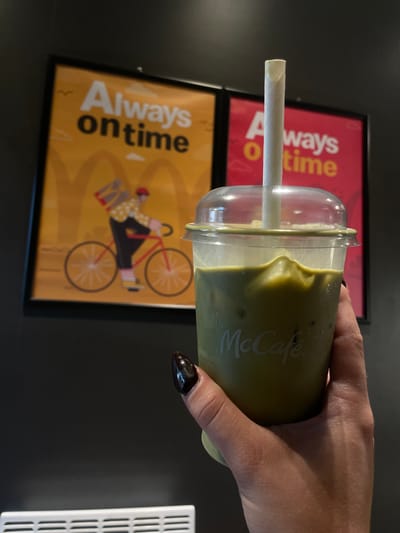 A hand holds a clear plastic McCafe cup filled with a light green beverage, likely a matcha latte, with ice. The cup has a white paper straw inserted. The McCafe logo is visible on the side of the cup in white lettering. In the background, two framed posters are partially visible. The upper poster has a red background with white and yellow text that reads "Always on time." The lower poster has a yellow background with black text that reads "Always on time" and features an illustration of a person riding a bicycle. The image appears to be taken indoors in a casual setting, possibly a cafe or a McDonald's restaurant. The lighting is artificial.