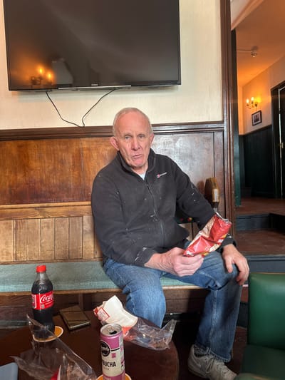 The scene depicts the interior of a public establishment, likely a pub or bar, located in Salisbury, United Kingdom. A Caucasian male, appearing middle-aged to elderly, with short grey hair and blue eyes, is seated on a green upholstered bench against a wooden paneled wall. He wears a dark grey Berghaus fleece jacket, stained with white paint, blue jeans, and light grey Nike athletic shoes. He holds a partially open red bag of crisps in his hands, which exhibit traces of white paint. His gaze is directed towards the viewer with a neutral expression.
On a dark wooden table in front of him are a partially full plastic bottle of Coca-Cola, a silver/gold-colored smartphone placed face down on a coaster, an open pink can of Momentum Kombucha (Cherry & Ginger flavor), several clear plastic wrappers, and a white paper wrapper. Above the man, a black Hitachi flat-screen television is mounted on a light-colored wall, with visible power cables. To the right of the man, an open doorway leads into a dimly lit area featuring a three-bulb wall-mounted candelabra and a framed picture on a darker paneled wall. A dark green upholstered chair armrest is visible to the right foreground.