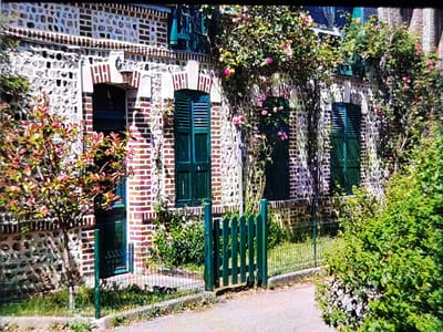 The image depicts a charming, stone and brick house with green shutters and a small green wooden gate. The architecture suggests a European style, possibly a historic building. The house is adorned with climbing pink roses, indicating a lush, garden setting. The surrounding vegetation is vibrant and green, with trees and bushes creating a natural, somewhat overgrown ambiance. The area appears to be a residential street or lane, with a paved surface in the foreground. The scene evokes a sense of rustic beauty and tranquility. The image is a photograph taken during the daytime, with natural lighting. The overall impression is one of a picturesque and well-maintained property in a scenic locale.