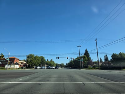 This image captures a view of an intersection in Elk Grove, United States of America, on a clear, sunny day. The sky is a vibrant, cloudless blue, suggesting it's likely daytime, possibly late morning or early afternoon. The scene is dominated by a wide, asphalt road with lane markings.
On the left side of the intersection, a building with an orange-tiled roof is visible, featuring signage that includes "Laguna" and a logo. Traffic lights are positioned at the intersection, with some showing red, indicating vehicles should stop. Several cars are present, stopped at or approaching the intersection, including white SUVs and a silver sedan.
To the right of the intersection, a street sign for "Elk Grove Blvd" is visible. Trees line the sides of the road, with lush green foliage. Power lines and utility poles are a prominent feature, stretching across the sky and along the roadside. The overall impression is one of a typical suburban or urban intersection, with everyday traffic and infrastructure. No people are clearly discernible in the image.