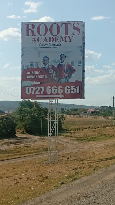 The image shows a large billboard advertising Roots Academy, a pre-school and primary school. "Learn to prosper" is the tagline. Two students in maroon sweaters stand in front of a school building on the sign. Below them, a red banner displays the phone number 0727 666 651 with an arrow pointing to the right. The billboard is supported by a white metal frame in a rural landscape with a dirt road. There are trees, shrubs and some buildings in the background under a sky with some clouds.