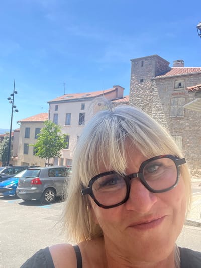 In Saint-Galmier, France, on a sunny day with a bright blue sky, a woman with blonde hair and dark-rimmed glasses takes a selfie. She is partially visible, with her face and upper chest in the foreground. Behind her, a street scene unfolds. Several cars are parked on the right side of the image, including a blue car and a grey Volkswagen Golf. The buildings in the background are made of stone and plaster, with terracotta roofs. A tall, stone tower with a small window stands out against the sky. A street lamp with multiple lights is visible on the left side of the frame. The overall impression is that of a quaint European town on a clear day.