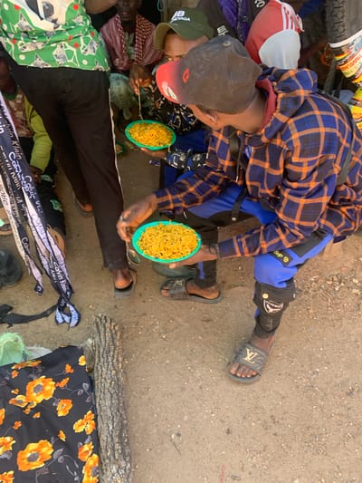 This media file depicts a group of people in what appears to be an outdoor, dusty setting, likely in Boi A, Nigeria. The subjects are clustered together on the ground, with some lying down and others seated or kneeling. One individual, wearing a blue and orange plaid shirt and blue pants with "Sup" and "Fashion" text, is holding a teal-colored bowl of noodles and a spoon, seemingly in the process of eating or offering the food. Another person, wearing a dark cap and a patterned garment, is also in close proximity to the food.

The scene suggests a moment of communal gathering, possibly a mealtime or a break. The ground is covered in dirt and small stones, indicating an unpaved area. Several individuals are covered by blankets or fabrics with various patterns, including a green and white geometric design and a black fabric with orange floral prints. Additionally, there are pieces of cloth with "Calvin Klein" text visible. The overall atmosphere appears informal and perhaps even a bit crowded. The lighting suggests it could be daytime, with natural light illuminating the scene. There are no strong indicators of specific weather conditions, but the dusty environment might imply dry weather.
