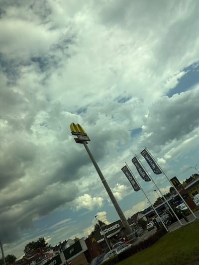 The image shows a McDonald's restaurant in Wieliczka, Poland. The sky is cloudy, with patches of blue visible. A tall sign with the McDonald's logo stands prominently, topped by the iconic golden arches and a McCafe sign. Several dark flags with the McDonald's logo are planted in a row, some of which are angled and appear to be blowing in the wind. In the background, the McDonald's building is visible, along with other structures and parked cars. A few people can be seen near the building and the drive-thru area. The scene appears to be set during daylight hours.