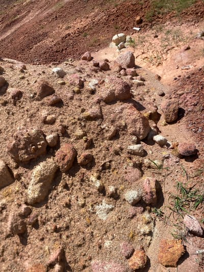 The image displays a close-up, ground-level view of a reddish-brown, sandy terrain with scattered rocks and sparse green vegetation. The landscape appears to be a hillside or embankment, with variations in soil composition and texture evident. Larger, rough-textured rocks are interspersed with finer sand and smaller pebbles. Patches of light-colored, possibly mineralized, deposits are visible on some of the rocks and in the soil. The lighting suggests a bright, sunny day, casting shadows that highlight the contours of the terrain. No people or discernible man-made structures are present. The scene is consistent with a natural, undeveloped area, possibly a construction site or erosion-prone zone.