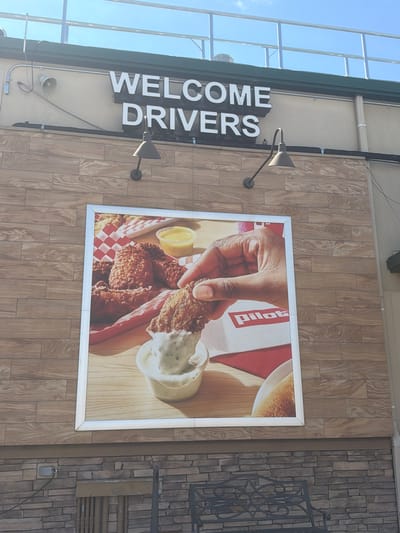 The image shows a sign that reads "WELCOME DRIVERS" above a large advertisement featuring fried chicken. The advertisement depicts a hand dipping a piece of fried chicken into a dipping sauce, with more chicken and a small cup of sauce in the background. The building has a facade of wood-like tiles, and below that, a stone facade. A metal bench is visible in the foreground. The overall scene appears to be an exterior view of a food establishment. The location is identified as the city of Lebanon, United States of America.