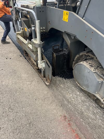 The image displays an active road paving operation in Dagonovo, Bulgaria. A large, grey asphalt paver machine, positioned on the right and center, is actively extruding and leveling fresh, dark black asphalt. A uniform strip of newly laid asphalt is visible to the left of the paver's screed. Further left and extending into the foreground, the existing light grey, granular road surface is present, featuring faint red spray paint markings in the lower right. The paver's tracked wheels are heavily coated with asphalt debris. Yellow warning decals, including symbols for 'beware of moving parts' and 'hearing protection,' are affixed to the machine's side panel. A clear plastic water bottle rests on the machine's upper surface. Partially visible on the far left, a worker wearing an orange high-visibility jacket, dark trousers, and work shoes stands on the newly paved asphalt, facing the operational machinery.