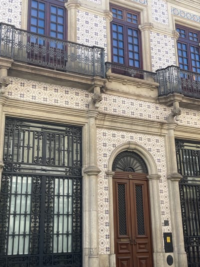 This media file is a photograph of the exterior of a building, likely in Águeda, Portugal, given the context. The building features intricate decorative tiling on its facade, with a repeating pattern of blue and white floral motifs. There are multiple windows visible, some with dark wooden frames and divided panes, and others protected by ornate black wrought-iron grilles.
The architecture suggests a historical or traditional style. A notable feature is a curved, elaborate dark wooden door or large window set within an arched stone surround, also decorated with tiling. There are also visible balconies with decorative metal railings. The overall impression is one of detailed craftsmanship and a rich aesthetic. The lighting suggests it is daytime, and the weather appears clear. No people are visible in the image. No discernible text is present in the image.