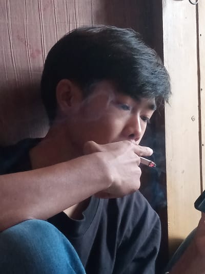 An indoor, close-up, slightly elevated perspective features an Asian male, wearing a dark short-sleeved t-shirt and blue jeans, seen from the chest up. He possesses dark hair and is looking downwards and to the right. He is actively smoking a lit cigarette, held between the index and middle fingers of his right hand, positioned near his mouth. The cigarette's tip glows orange-red, and visible white smoke rises around his face. The background consists of vertical wooden paneling: a reddish-brown section with visible grain and possible staining occupies the left, transitioning to a lighter, pale yellow/beige wooden panel or frame on the right. An indistinct dark object is partially visible in the bottom right foreground. The specific location within Muka, Indonesia, cannot be determined from the visual evidence presented in the image.