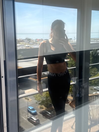 A woman is taking a mirror selfie with her phone on a balcony. She is wearing a black sports bra and black leggings. Her abdomen is visible and appears toned. The background shows a city view with buildings, trees, and cars. The image is taken from a reflection in a glass window, likely from a building in Loures, Portugal.