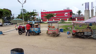This is an outdoor daytime, eye-level, full shot of a street scene with several food carts and a building in the background. The ground is unpaved and sandy, with some gravel and patches of dry grass. A large red building with white trim and windows is visible in the background. Several flags are flying from the building. To the left of the building, there are trees and utility poles with wires.
In the foreground, there are three food carts. The leftmost cart is blue and appears to be a "Baksos Goreng" (fried meatballs) cart. A person in a red shirt and dark pants is standing next to it, interacting with the cart. Behind this cart is a motorcycle, also with a blue top cover. To the right of the blue cart is a pink and red cart labeled "BAKSO CRISPY" and "BATAGOR & SEMPOL AYAM." This cart has an umbrella providing shade. Further to the right, there is another cart with a purple umbrella, displaying various snacks and drinks. A rack of small packaged items is positioned in front of this cart.
On the road in the background, a white SUV is visible, along with two people walking. A motorcycle is also seen in the distance. The sky is overcast with some clouds. The scene appears to be in a relatively open area, possibly a marketplace or a roadside location. The overall impression is one of informal street food vending.