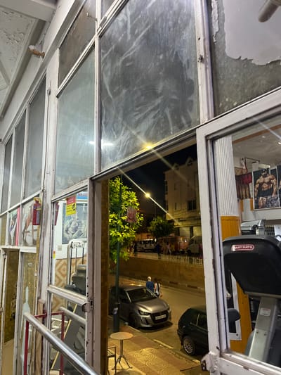 The image shows a view through large, grimy windows, likely from inside a gym or fitness center, looking out onto a street in El Hajeb, Morocco at night. Peeling paint is visible on the window frames and the ceiling. Inside, posters of muscular bodybuilders adorn the walls. Various gym equipment, including what appears to be a treadmill, is present. A red fire extinguisher is mounted on the wall. Outside, a tree with bright green foliage is illuminated by streetlights. Cars are parked on the street, and a few people are walking on the sidewalk. The street is paved with yellow and beige patterned tiles. The buildings across the street are lit up, suggesting a commercial or residential area. The overall impression is one of a somewhat dated and well-used establishment.