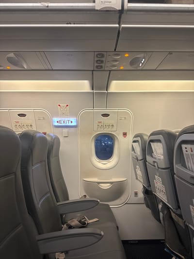 The image depicts the interior of an airplane cabin. Emergency exit row seats are visible, located next to an emergency exit door with a window. The exit sign is illuminated. Overhead panels with passenger controls and ventilation are visible. The location is not Omoecheigbo, Nigeria.