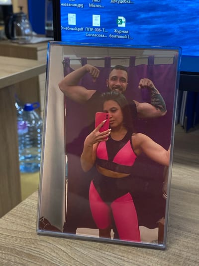 A photograph in a plastic frame rests on a wooden surface, possibly a table. The photo within the frame shows a couple flexing their muscles while posing for a selfie. The woman is holding a pink smartphone. The man has tattoos on his left arm. The woman is wearing a pink and black sports bra and matching leggings. The setting is indoors, with a computer monitor visible behind the photo, displaying Russian text. A water bottle sits on the table as well. The location and city, Luzhitsy, Russia, cannot be determined from the image alone.