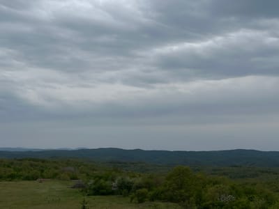 The image captures a landscape near Gramatikovo, Bulgaria under a cloudy sky. The foreground consists of a grassy field with some sparse vegetation and what appears to be a small clearing with two structures of unknown function. The middle ground is dominated by rolling hills covered in dense forest. In the distance, more hills are visible, their outlines blurred by haze. The sky is overcast with thick, gray clouds covering the entire upper half of the image. There are no people or animals visible.
