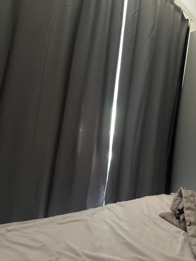 A low-angle shot shows a partial view of a room. To the left, heavy, dark gray curtains hang, allowing a horizontal sliver of bright light to penetrate through a gap. To the right, a light gray, possibly white, duvet or comforter is partially visible, with a section folded over at the top. The scene is simply a view of these items, without any apparent people, actions, or interactions taking place. The location Бобровский, Russia is not discernable from the details in this image.