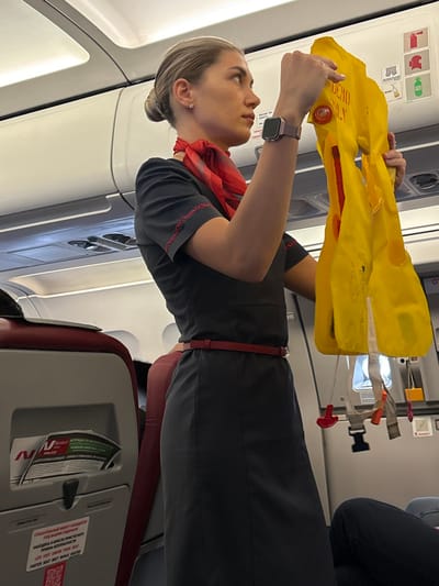 Inside a commercial aircraft cabin, a female flight attendant with blonde hair in a bun wears a dark grey short-sleeved uniform dress with red embroidery, a red neck scarf, and a red belt. A purple-banded smartwatch is visible on her left wrist. She holds a yellow life vest marked "DEMO ONLY" with both hands, presenting it. Her gaze is directed upwards and to the right. The background shows white overhead bins, various safety instruction decals (fire extinguisher, oxygen mask, life vest, green exit signs), and the top of a red-upholstered passenger seat. A safety information card, partially in Russian, is in the seatback pocket, and text below it includes "FASTEN SEAT BELT WHILE SEATED." The aircraft is situated in the city of Novogorelovo, Russia.