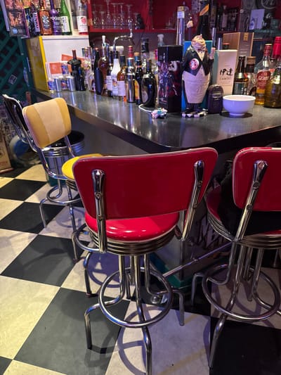 The image shows a bar setting with multiple red and chrome bar stools arranged around a counter. Behind the counter, shelves are stocked with various bottles of alcohol, glassware, and other bar-related items. The floor is a black and white checkerboard pattern.
One bar stool in the foreground has a light tan seat and a red back. Another bar stool behind it is entirely red with chrome accents. A third bar stool, mostly obscured, appears to be yellow.
On the counter, there are several bottles of alcohol, some in decorative packaging. One item on the counter resembles a small white bowl. There is also a figure of a man in a suit on the counter.
The lighting in the scene is dim, suggesting it is nighttime or the bar is intentionally lit this way. The reflections on the counter indicate a glossy surface.
There is some text visible on the bottles and packaging behind the counter, but it is not clearly legible in most cases. Some visible text includes "JINRO" on a bottle, and parts of other labels that are too small or blurred to read. The overall scene suggests a vibrant and perhaps retro-themed bar environment, possibly in Adachi, Japan, as suggested by the location context. No people are visible in the image.