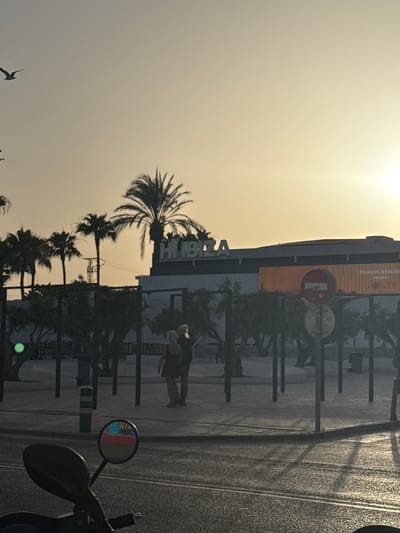 The image depicts a street scene in Platja D'en Bossa, Spain, during what appears to be sunset or early evening. A building with the illuminated word "HÏ" and part of "BÖŻA" is visible in the background, suggesting it is the Hï Ibiza nightclub. Palm trees are silhouetted against the bright, hazy sky. Two individuals, a man and a woman, are standing and facing each other on a paved area with distinct shadows cast by the setting sun. In the foreground, the handlebars and side mirror of a motorcycle are visible. The mirror reflects colored lights, possibly from a traffic signal or nearby signage, and the overall lighting suggests a warm, low-light environment. The street surface is wet, indicating recent rain or cleaning, and is marked with white lines. A traffic sign with a red circle and horizontal bar, indicating "no entry" or a similar prohibition, is mounted on a pole. Another smaller circular sign is below it. A green circular light is visible on a pole to the left of the frame.