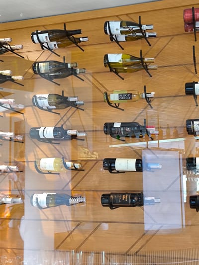 The image shows a wall-mounted wine rack displaying various bottles of wine. The rack is constructed with black metal supports that cradle each bottle horizontally. The backdrop appears to be a light-colored wooden panel with geometric patterns, possibly a decorative element of the interior. Some of the bottles are clearly labeled, revealing the brands, types, and names of the wine. The wines visible include SCHIEF ER, PINOT GRIGIO, MAJANIU, 90 MOUTON, MASCOTA, and Casillero Diablo. The bottles are placed strategically to show their labels. The background shows objects through the glass, which may include a wine rack. There is no other visual information in the scene to suggest its location in Sidemen, Indonesia.